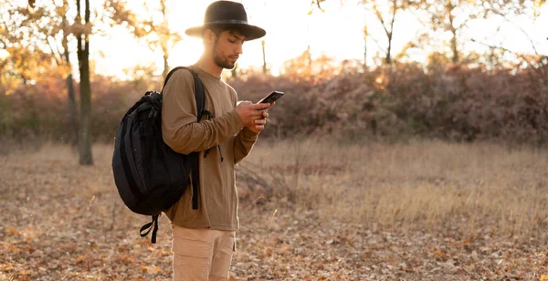 A traveler in a hat catches a connection and the Internet while standing in the forest at sunset. Autumn hiking in the mountains. Copy space