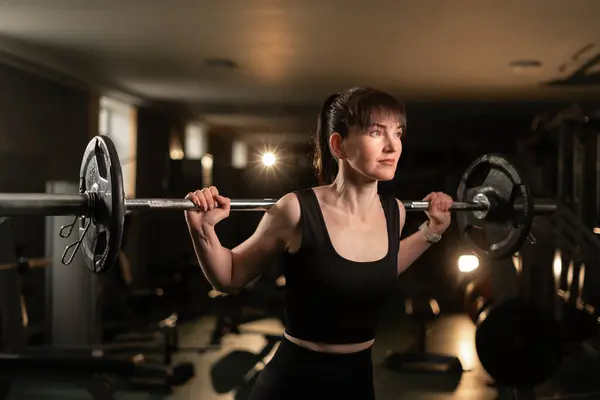 Woman doing squats using barbell. Concentrated fitness woman with muscular body training in gym in dark smoky atmosphere. Copy space