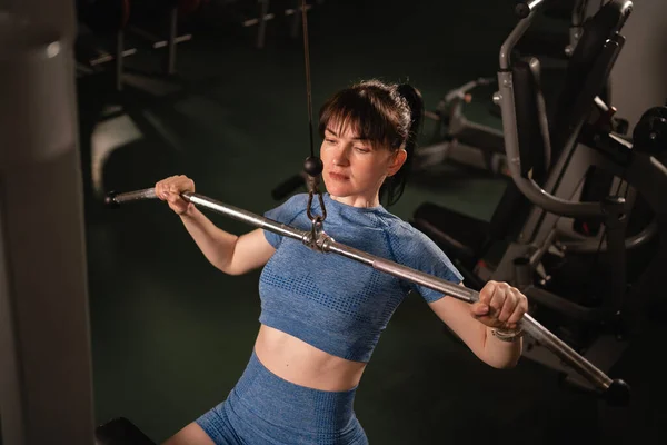 young beautiful fitness lady in blue sportswear sits on a simulator and performs exercises for arms and back in gym. A woman with a muscular body is exercising. Sport and fitness concept