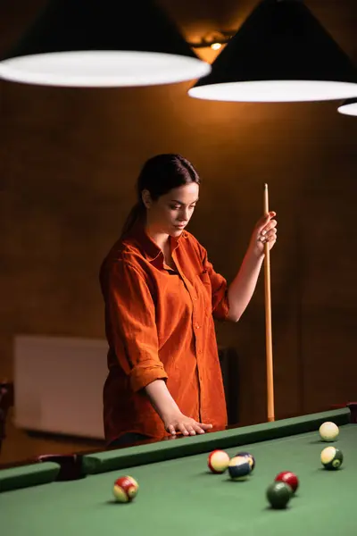 A young woman playing billiards stands thoughtfully near the billiard table with a cue in her hands. Copy space