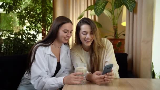 2人の幸せな若い女性が屋内のカフェでスマートフォンでソーシャルメディアを閲覧しています ライフスタイル 人々のコンセプト — ストック動画