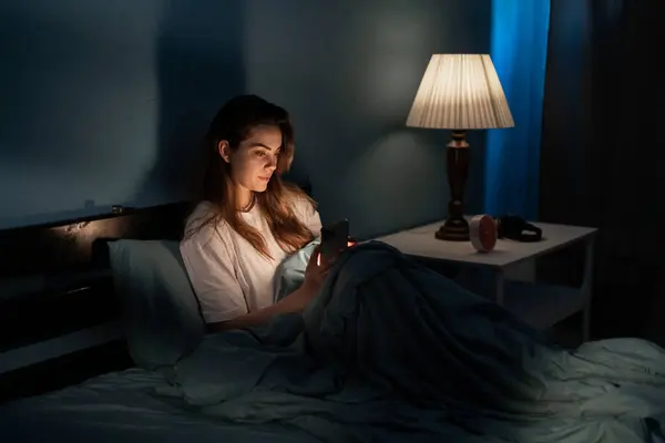 Attractive woman uses smartphone in bed at home at night. Browsing social media, reading news at night. Copy space