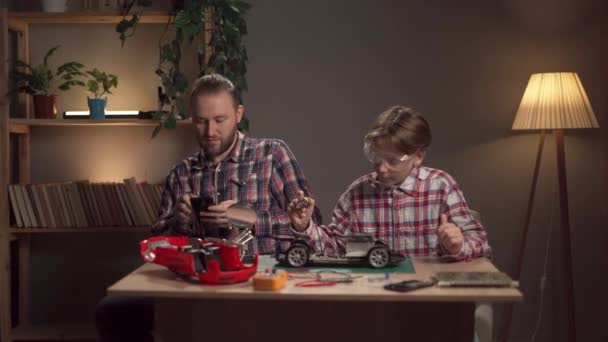 ハッピーな父親は 自宅でリモートコントロールされた車をはんだ付けするために息子に教えています 壊れたおもちゃを修理する男と少年 コピースペース — ストック動画