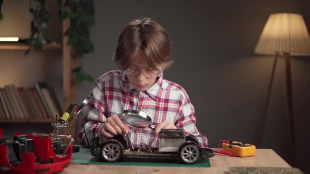 可爱的男孩与放大镜坐在桌旁与破碎的玩具车 修理的概念 复制空间 — 图库视频影像