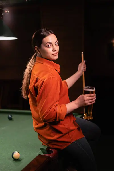 Young Caucasian woman playing billiards in a bar, drinking beer and holding a cue. Looking at camera and smiling