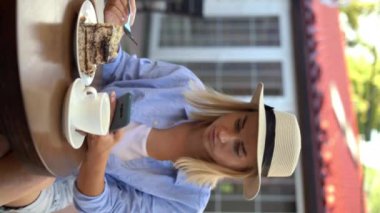 Bir sokak kafesinde hasır şapkalı güzel sarışın kız kahve içer ve sosyal ağlara göz atarken ya da mesajları okurken cep telefonu kullanır. Dikey video