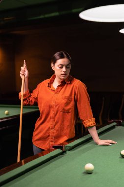 Güzel bir kadın elinde ıstakayla bilardo masasının yanında duruyor ve bilardo maçına hazırlanıyor. Boşluğu kopyala