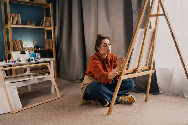 Concentrated Caucasian woman artist sitting on floor in studio painting drawing on canvas, creativity work. Copy space