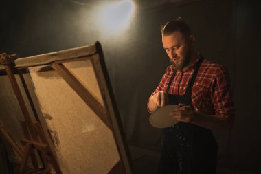 Erkek sanatçı, karanlık bir sanat stüdyosunda resim yaparak yağlı boyalarla bir başyapıt yaratıyor. Boşluğu kopyala