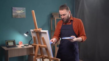 Yakışıklı bir adam tuvale yağlı boyalarla resim çizer. Boşluğu kopyala