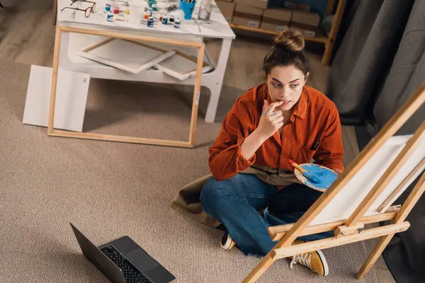 Talented Serious woman professional artist sitting on floor in her studio painting drawing on canvas, creativity work, contemporary creative painter. Copy space