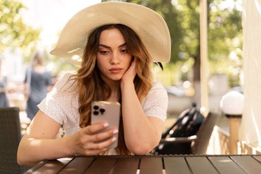 Hasır şapkalı şık esmer kadın kahve dükkanında dinlenirken cep telefonuyla iyi haberler okuyor, boş zamanlarında kafede dinlenirken cep telefonuyla konuşan kadın. Boşluğu kopyala