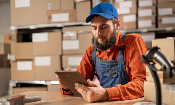 A warehouse worker conducting an inventory uses a digital tablet while sitting at a table in work clothes. Copy space
