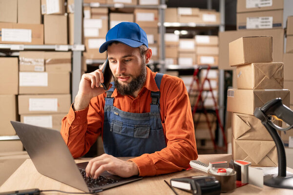 Male entrepreneur speaking on the phone while working on a laptop in warehouse. Online store owner making product shipping. E-commerce concept