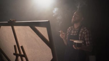 Geniş bir stüdyoda sigara içen Millennial bir adam büyük bir tuvalle modern sanatın bir başyapıtını resmediyor. Karanlık sanat atölyesi. Boşluğu kopyala