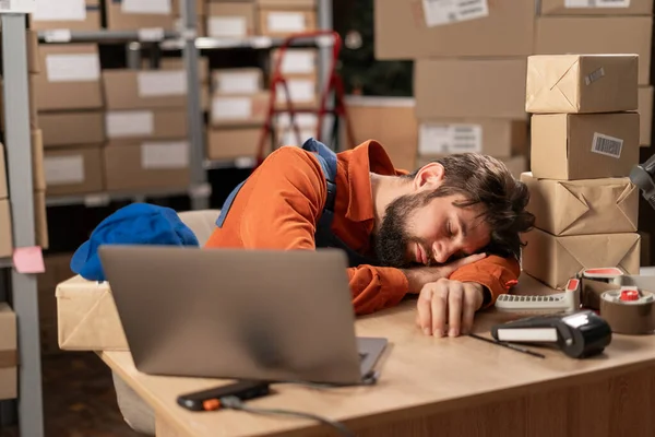 Exhausted man sleeping on his desk near laptop tired of overworking in warehouse. Workaholic suffering from chronic fatigue. Copy space