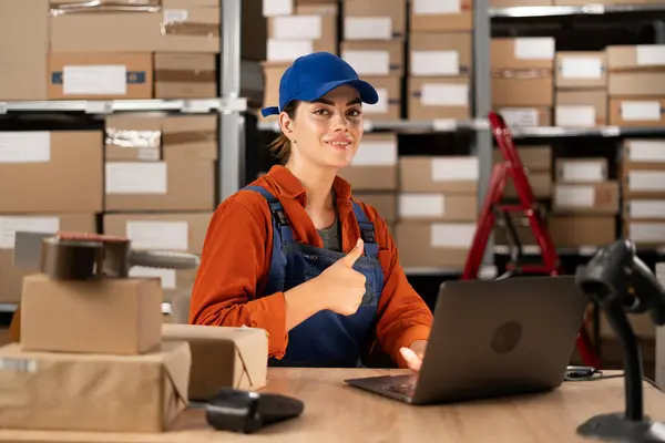 Manager checking online orders and inventory on a laptop while sitting at a desk working in warehouse with shelves full of parcels, showing thumb-up. Copy space