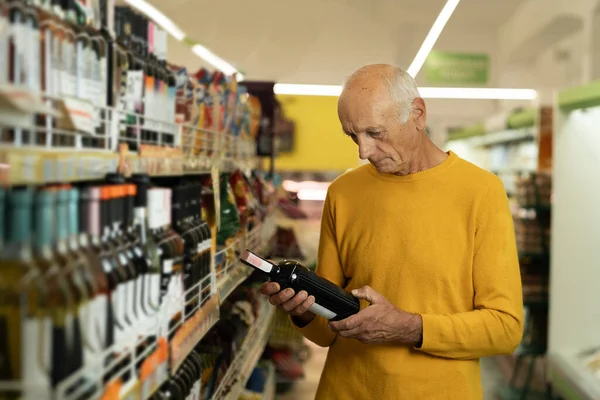 Elderly man chooses wine in the store. Senior man reads the label on a bottle of wine before buying. Copy space