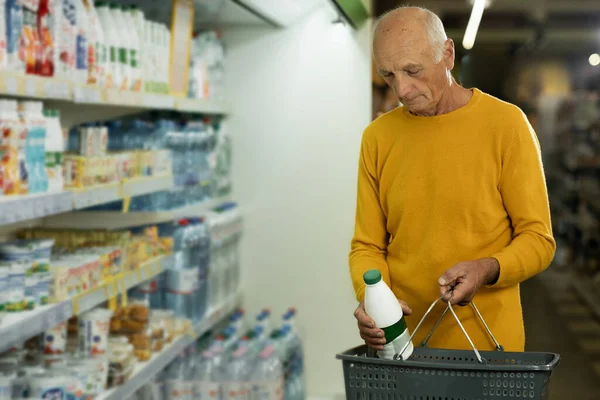 Senior man holding milk bottle and shopping basket choosing food on shelf with dairy products. Grocery shopping concept
