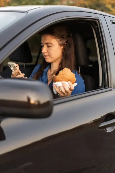 A woman eating a fresh hamburger while driving a car uses a smartphone to communicate online. Multitasking concept