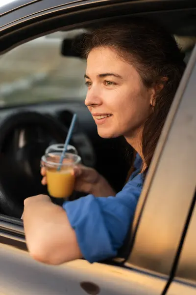Happy young woman with cocktail driving her car. Woman sipping a cold juice while in car. Young driver drinking beverage. Copy space
