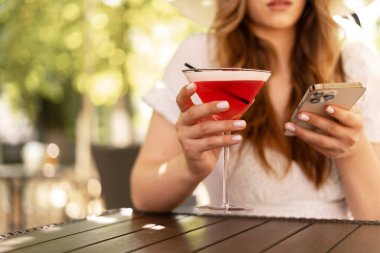 Genç şık bir kadın kokteyl içiyor, yazın kafede dinleniyor, sosyal ağlarda sohbet etmek için cep telefonu kullanıyor. Tatil ve yaz tatili konsepti. Yakın plan.