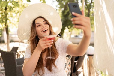 Büyük yaz hasır şapkalı güzel bir kız güneşli terasta selfie çekiyor, bir kafede kokteyl içiyor. Boşluğu kopyala
