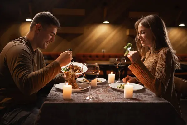 young couple in love having dinner in a restaurant celebrating Valentine's Day, man putting pasta in a plate and woman smiling. Romantic dinner concept