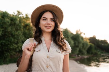Seyahat konsepti. Yaz tatilinde şapka takan ve kameraya bakan sevimli bir kadının portresi. Kumsalda yürüyen bir turist. Boşluğu kopyala