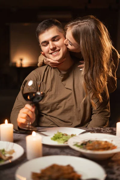 Happy young couple in love drinking wine, wife hugging and kissing her husband, celebrating Valentine\'s day having romantic dinner with candles at home. Copy space