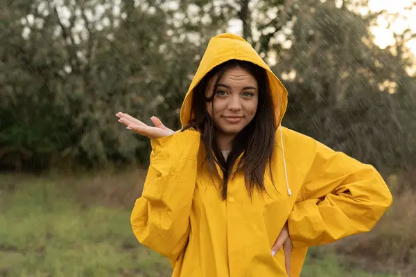 Young woman wearing yellow raincoat enjoying the rain outdoors in the park, catching the rain drop with hand. Copy space