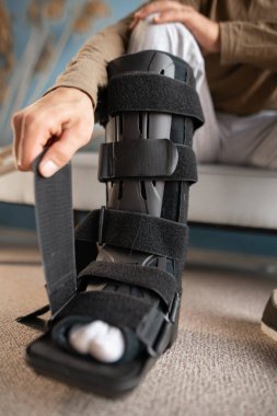 Genç adam evdeki kanepede otururken bağları desteklemek için bacağına ayak bileği ortozu takıyor. Tıp konsepti. Yakın plan.