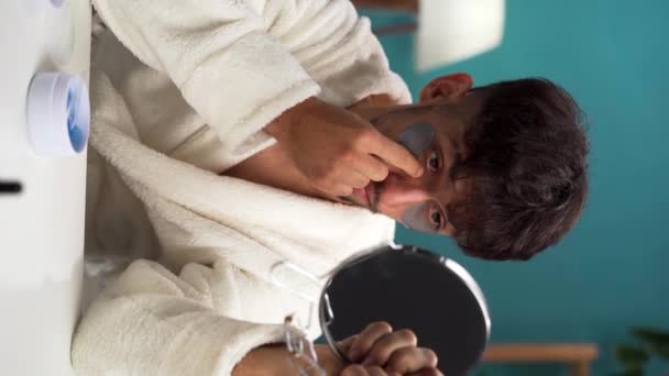 年轻英俊的男子穿着浴袍从家里的脸上摘下胶原蛋白眼斑 每天早晨的美容程序 男性恐慌和年轻化的概念 垂直录像 — 图库视频影像