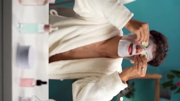 英俊的年轻人 脸上戴着面具 把一片新鲜黄瓜放在眼睛上 皮肤护理 治疗和美容概念 垂直录像 — 图库视频影像