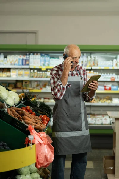 Old male sales assistant in a grocery using digital tablet and talking on cell phone. Owner with digital tablet preparing online grocery order for his shop. Copy space
