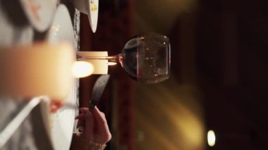 Genç çift restoranda akşam yemeği yiyor kırmızı şarap bardaklarını tokuştururken masada mumlar ve tabaklar eşliğinde sevgililer gününü veya evde yıldönümünü kutluyorlar. Dikey video