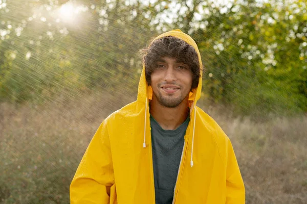 Arabic young man wearing a yellow raincoat during the rain in park. Happy guy enjoying the rain outdoors, has a joyful expression in the rainy weather. Copy space