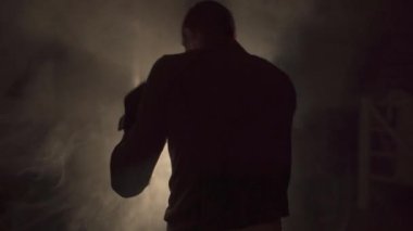 Spor salonundaki dumanlı arka planda boks eldivenleriyle torbaya yumruk atan genç bir adam. Düşük açı. Boşluğu kopyala