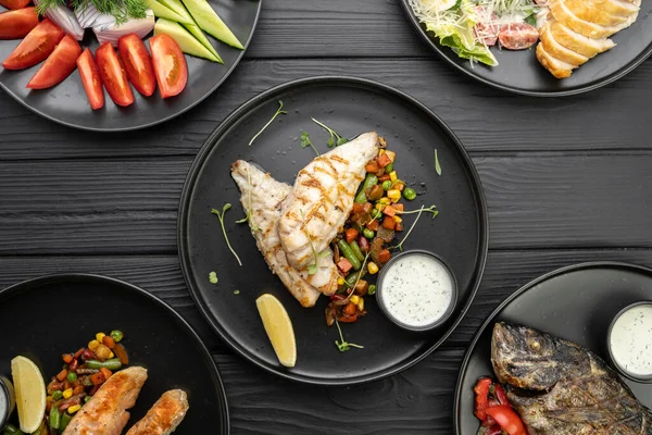 Top view of various grilled dishes served on a dark table in a restaurant. Fish fillet with vegetables. Flat lay