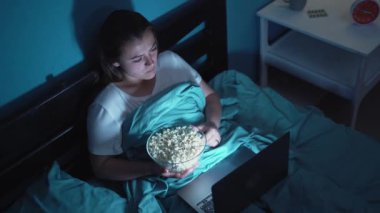 Çekici genç bir kadın battaniyenin altında yatıp dizüstü bilgisayarında film izlerken patlamış mısır yiyor. Boşluğu kopyala