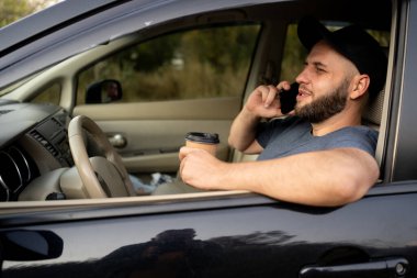 Genç sakallı adam bir arabanın direksiyonunda oturup kahve içerken telefonla konuşuyor. Beklerken park yeri trafiği sıkışık. Boşluğu kopyala
