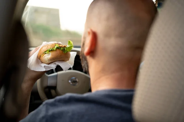 Man driving car while eating hamburger, back view. Waiting and standing in traffic jam. Copy space