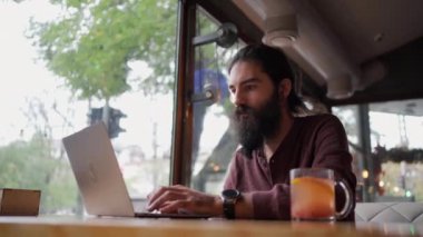Uzun saçlı, sakallı, beyaz bir adam bir kafede oturup dizüstü bilgisayar başında çalışıyor. Serbest çalışan kavramı kaldırıldı. Boşluğu kopyala.