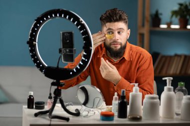 Sakallı adam güzellik vlogcusu. Sosyal medyada akıllı telefon paylaşımı videosu. Erkek blogcu makyöz ya da kozmetikçi yaşıyor. Cilt bakımı rutin dersleri kaydediliyor. Boşluğu kopyala