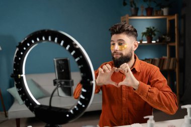 Sakallı adam, güzellik vlogcusu, kalp hareketi gösteriyor. Sosyal medyada akıllı telefon paylaşımı videosu. Erkek blogcu makyöz ya da kozmetikçi yaşıyor. Cilt bakımı rutin dersi. Boşluğu kopyala