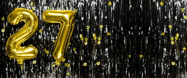 Gold foil balloon number number 27 on a background of black tinsel decoration. Birthday greeting card, inscription twenty seven. Anniversary event. Banner. copy space.