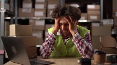 Bilgisayar işinden yorgun düşen yorgun genç adam, baş ağrısı çeken adam dizüstü bilgisayar kullanarak depoda oturuyor. Boşluğu kopyala