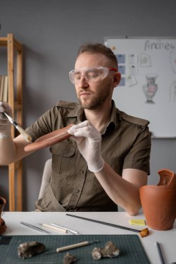 Erkek arkeolog ofiste çalışırken tozdan antika bir plakayı fırçayla temizliyor. Boşluğu kopyala