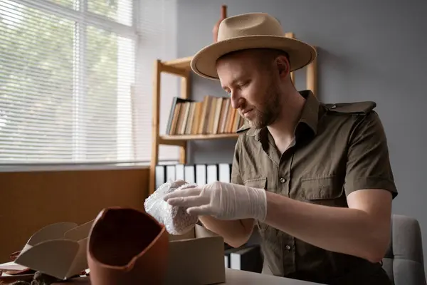 Archaeologist or digger in a hat working in an office, packing antique dishes for sale. Copy space