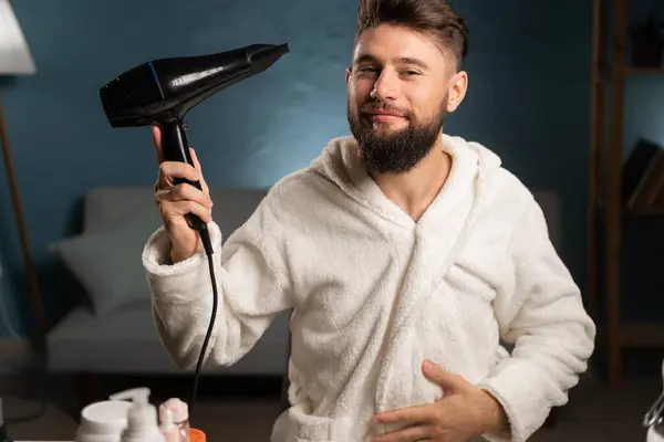 Young man drying and styling hair using hairdryer at home after shower. Hair care and hairstyling concept. Male beauty routine. Copy space
